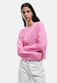 Suéter de punto rosa ligero y acanalado con mangas anchas, que presenta un escote redondo. Combinado con jeans blancos de tiro alto.