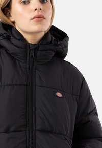Black puffer jacket with a high collar, featuring a full zipper and a small logo patch. Texture is smooth with quilted sections.
