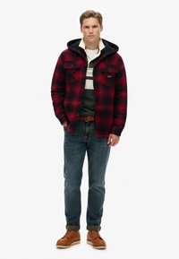Rød og sort ternet jakke med hætte, to frontlommer, lagdelt over en stribet skjorte og blå jeans, parret med brune lædersko.