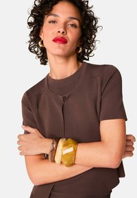 Bruine gebreide top met korte mouwen en een ronde halslijn, met een metalen sluitingdetail. Multikleurige chunky hars armbanden om de arm.