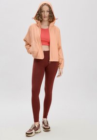 Leichter pfirsichfarbener Zip-Hoodie mit Kordelzugkapuze, getragen über einem roten Croptop und burgunderfarbenen Leggings, kombiniert mit braunen Sneakern.