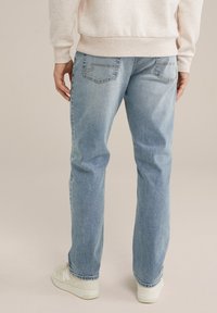 Šviesiai mėlyni džinsai su tiesia pėdos forma, turintys užpakalines kišenes ir minimalų blukimą, derinami su kreminės spalvos kedais.