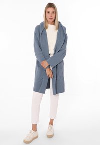Jasnoniebieski, oversize kardigan z kapturem, o fakturowanej tkaninie, otwarty z przodu, z dwiema bocznymi kieszeniami, w połączeniu z białymi spodniami i jasnymi butami.