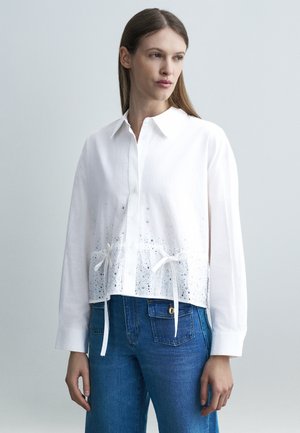 Žena v bielej košeli s gombíkmi, ozdobenej dierkami a stužkami na spodnom okraji, v kombinácii s modrými rifľami, otočená doľava.