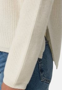 Pull en maille de couleur blanc cassé, présentant une texture côtelée et des manches ajustées. Associé à un jean en denim bleu, mettant en avant un design taille haute.
