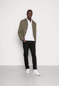 Olivengrønn lett jakke med knappelukking, hvit poloskjorte, svarte slim-fit bukser og hvite joggesko. Ensfarget bakgrunn.
