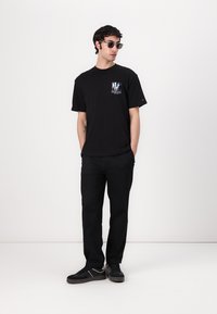 Zwarte katoenen T-shirt met een multicultureel logo op de linkerborst, gecombineerd met zwarte broek en zwarte sneakers. Model draagt een zonnebril.