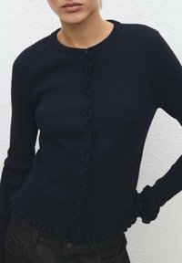 Hladký námořnický svetr s kulatým výstřihem, s volánkovými okraji podél knoflíkové légy a na rukávech, má přední knoflíky a úzký střih.