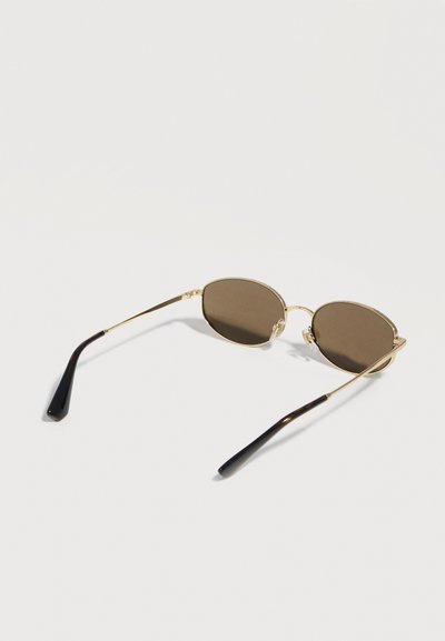 Οβάλ γυαλιά ηλίου με χρυσή κορνίζα, σκοτεινούς φακούς και μαύρα ακροωτάρια, που ακουμπούν σε λευκή επιφάνεια.