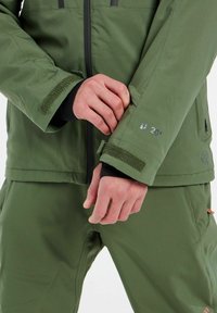 Grøn vandtæt jakke med sorte detaljer, der har justerbare manchetter og et synligt logo med teksten "20K" påærmet.