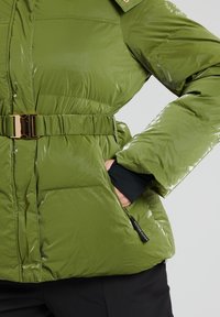 Grön puffy-jacka med en glänsande yta, bälte i midjan och guldiga detaljer. Har fickor och hög krage, designad för värme och komfort.
