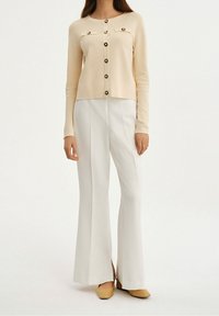 Beżowy kardigan z dwoma przednimi kieszeniami i czarnymi guzikami, w połączeniu z białymi spodniami w fasonie dzwonów i żółtymi butami na obcasie. Gładka tekstura.