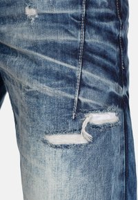 Jean en denim bleu clair avec un design déchiré, présentant des déchirures effilochées et un dégradé de couleur, ainsi que des coutures contrastantes le long des coutures.