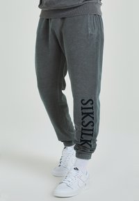 Šedé tepláky vyrobené z měkké tkaniny, se bočními kapsami a černým logem na levé nohavici. Spárovány s bílými teniskami.