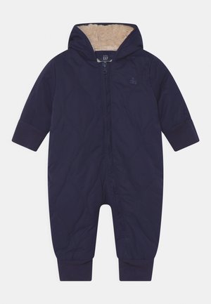 Marineblauwe gewatteerde simpele overall met een capuchon, voorzien van een zachte, beige fleecevoering en elastische manchetten. Voorkant met ritssluiting en reliëf logo-detaillering.