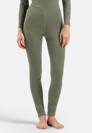 Person, die olivgrüne, enganliegende Rippleggings und ein passendes langärmliges Oberteil trägt, barfuß vor einem weißen Hintergrund steht.