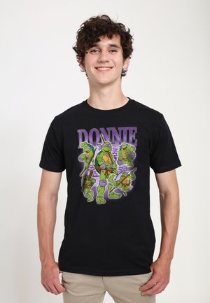 Jeune homme portant un t-shirt noir avec un motif Donnie des Tortues Ninja, souriant, debout devant un fond blanc uni.