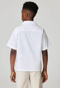 Camicia bianca in cotone a maniche corte con colletto classico, caratterizzata da una vestibilità rilassata e dettagli con spacchi laterali, indossata con pantaloni beige.