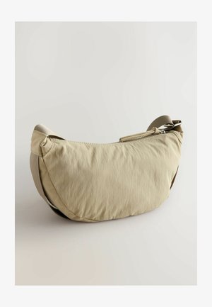 Beige kangaskassi, jossa on kaareva muoto, hienovarainen tekstuuri ja vetoketjullinen pää. Varustettu sävystä säädettävällä hihnalla kantamista varten.