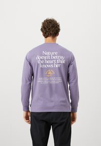 Long-sleeve, purple cotton shirt featuring white and gold text on the back. Text includes a quote and brand logo, with ribbed cuffs.