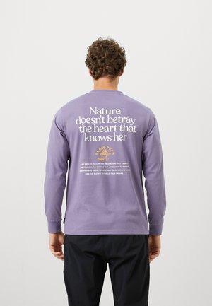 Chemise en coton à manches longues, de couleur violette, avec un texte blanc et doré au dos. Le texte comprend une citation et le logo de la marque, avec des poignets côtelés.