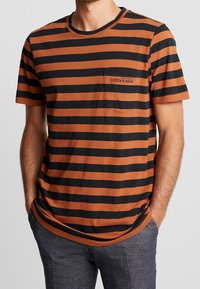 Hombre que lleva una camiseta de manga corta a rayas horizontales en marrón y negro con un bolsillo en el pecho y pantalones gris oscuro.