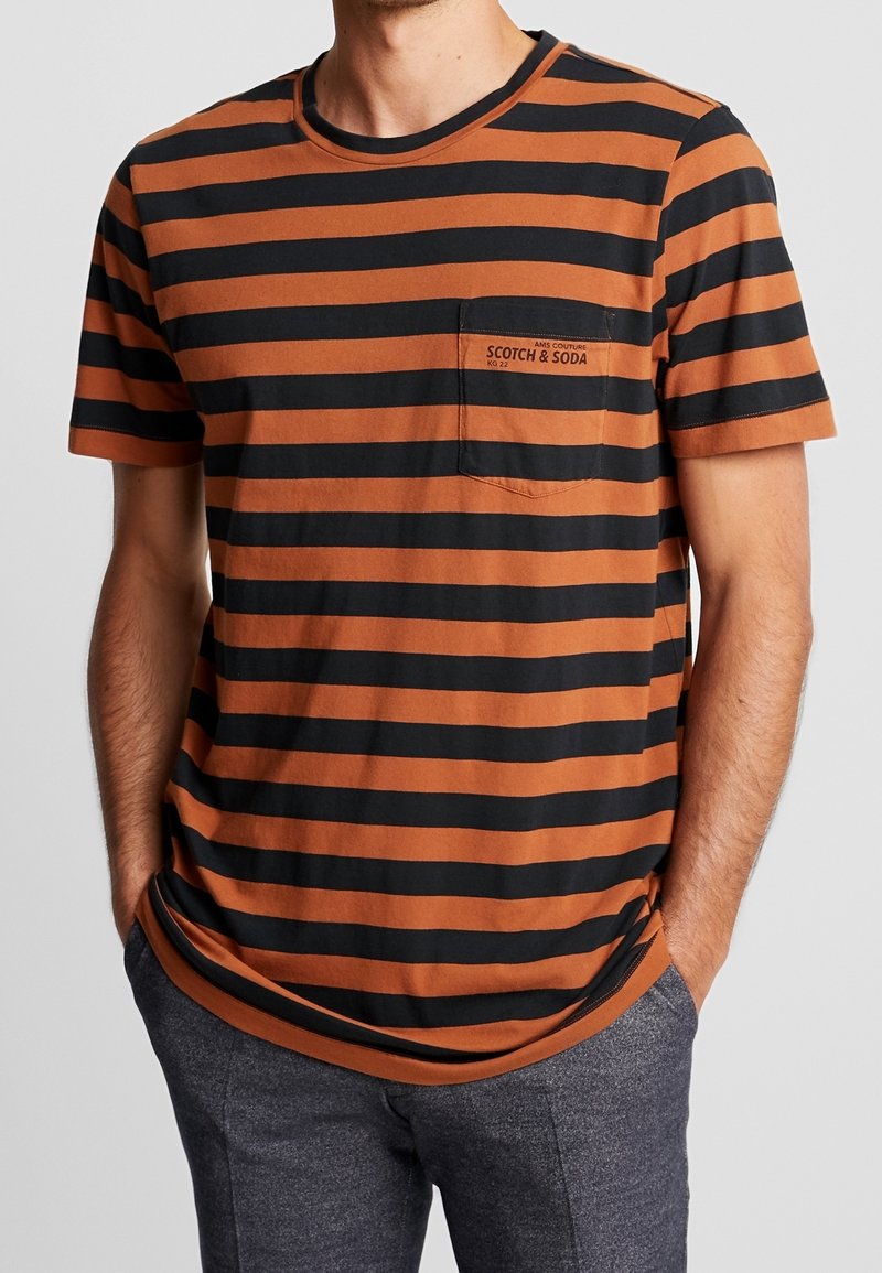 Hombre que lleva una camiseta de manga corta a rayas horizontales en marrón y negro con un bolsillo en el pecho y pantalones gris oscuro.