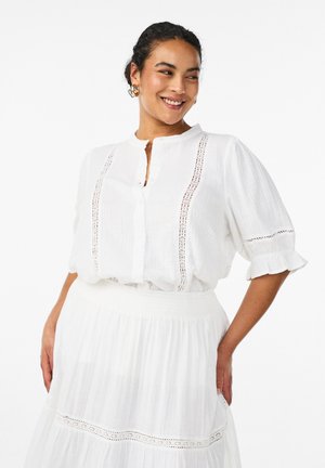 Usmievavá žena v bielej blúzke s čipkovými detailmi a zodpovedajúcou sukňou, ruky na bokoch, pozerá sa do strany na jednolatom pozadí.