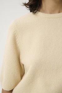 Short-sleeved, cream sweater made of soft, textured fabric. Features a ribbed neckline and a relaxed fit with subtle construction details.