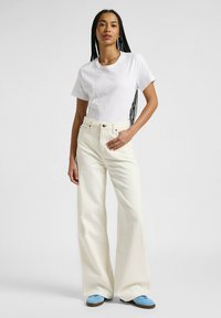 Witte T-shirt met korte mouwen, crèmekleurige hoge taille wijde jeans, blauwe schoenen met bruine rubberen zolen. Subtiele textuur en strakke lijnen.
