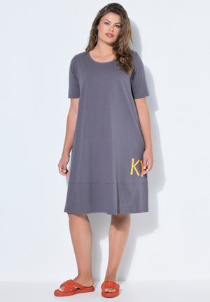 Kobieta o falowanych włosach, ubrana w luźną, sięgającą kolan szarą sukienkę z krótkimi rękawami i pomarańczowe wsuwane sandały, stoi na tle białego tła.