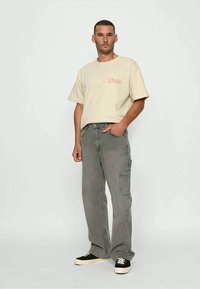 Man står klädd i en beige överdimensionerad t-shirt med röd text, grå cargojins och svarta låga sneakers med vita sulor.