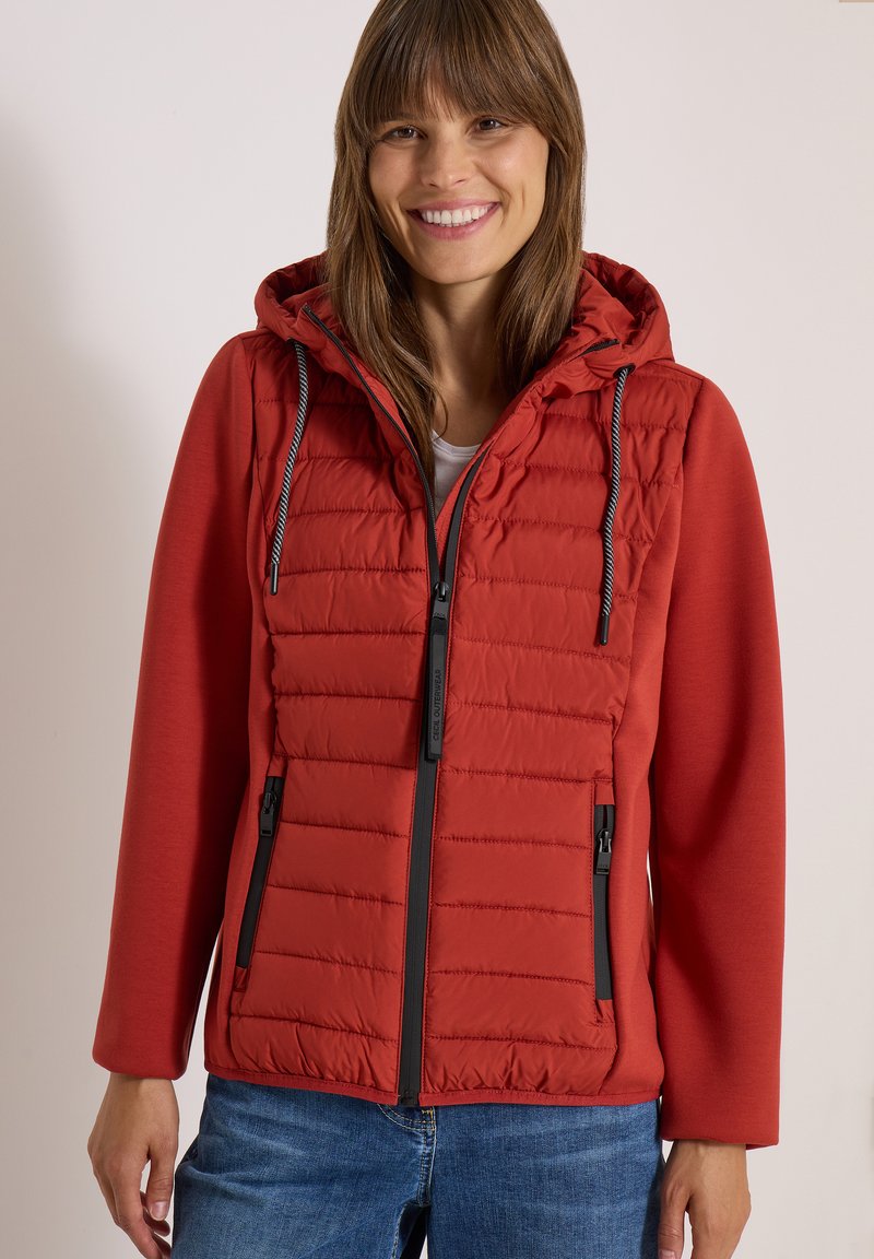 Femme souriante portant une doudoune rouge à capuche avec des poches zippées et un jean bleu sur un fond uni.