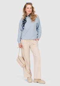 Felpa azzurra chiara con scollatura rotonda, pantaloni beige a gamba larga e borsa a tracolla beige. Foulard a righe rosse e blu come accessorio.