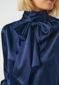 Blusa in satin blu navy con colletto alto e ampio fiocco al collo; texture liscia e leggero lucido; vestibilità aderente con spalle arricciate.