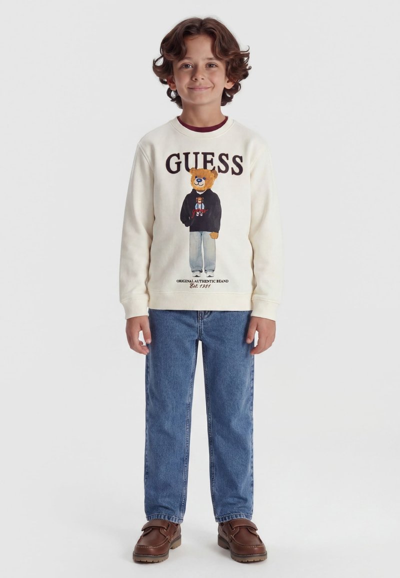Enfant aux cheveux bouclés porte un sweat-shirt blanc avec un ours en peluche et le texte "GUESS", un jean bleu et des chaussures marron, debout devant un fond uni.