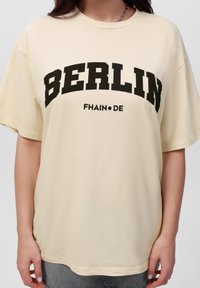 Kremhvit bomulls T-skjorte med oversized passform, med fett svart tekst som leser "BERLIN" og "FHAIN•DE" over brystet. Korte ermer.