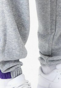 Pantaloni grigi in felpa con polsini elasticizzati, caratterizzati da un accento viola alla caviglia. Abbinati a sneaker bianche, che mostrano un tessuto texturizzato.