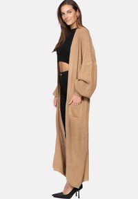 Lång stickad cardigan i beige med vida ärmar, öppen front, ribbad textur och två framfickor, parat med en svart topp och byxor.