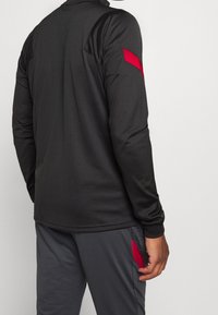 Sort langærmet træningsshirt med røde detaljer på skuldrene, parret med grå bukser med røde sidelommer. Glat, letvægtsstof.