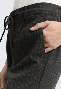 Zwarte pinstripe broek met een zachte textuur, voorzien van een koord in de taille en zijzakken, gepresenteerd tegen een lichte achtergrond.