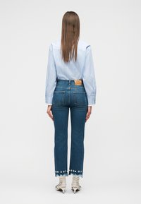 Mujer de pie de espaldas, vistiendo una camisa de rayas azul claro, vaqueros cortos con dobladillo floral y sandalias blancas con tacón.