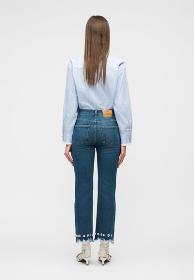 Γυναίκα που στέκεται με την πλάτη, φοράει γαλάζιο ριγέ πουκάμισο, κοντά τζιν με φλοράλ τελείωμα και λευκά πέδιλα με τακούνι.