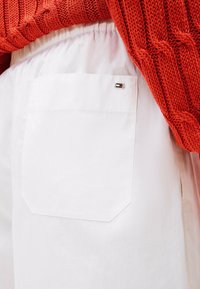 Pantaloni bianchi con una tasca posteriore visibile e un piccolo dettaglio logo colorato. La tasca ha un fondo arrotondato ed è cucita al tessuto.