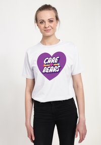 Camiseta blanca de algodón con un gran gráfico de corazón morado que tiene el texto "CARE BEARS" y elementos decorativos coloridos dentro del corazón.