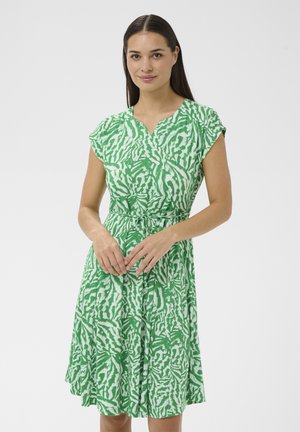 Mujer con cabello largo y oscuro que lleva un vestido de longitud hasta la rodilla con un patrón verde y blanco, mangas cortas y un lazo en la cintura.