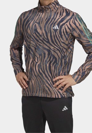 Homme portant un haut de sport à manches longues marron et noir à rayures zèbre avec une fermeture éclair au quart et un pantalon noir avec un logo blanc.