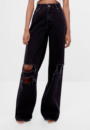 Schwarze High-Waist Wide-Leg-Jeans mit großen horizontalen Rissen an beiden Knien, getragen von einer stehenden Person mit offenen Schuhen.