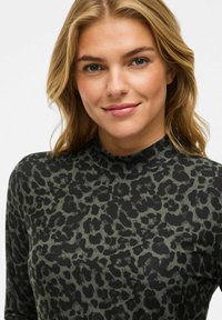 Jonge vrouw met blond haar die een zwart en donkergroen luipaardprint hoge hals top draagt, glimlachend tegen een effen achtergrond.