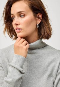 Maglione grigio a collo alto in morbido tessuto con polsini a coste. Il modello ha un taglio di capelli a caschetto corto e indossa orecchini tondi in argento.